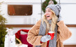 © Prostock-studio - A joyful woman wearing a warm coat, hat, and scarf is sipping from a red mug in a winter setting, surrounded by holiday decorations and a festive atmosphere.