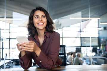 Wall Mural - Portrait of young dreaming Indian business woman manager ceo using cell phone mobile app. Pondering Latin Hispanic female businesswoman holding smartphone in office for trading, reading, work online