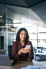 Canvas Print - Young Indian business woman manager ceo using cell phone mobile app laptop. Smiling Latin Hispanic mature female businesswoman holding smartphone in office trading, reading, working online. Vertical