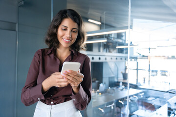 Canvas Print - Happy young Latin Hispanic business woman holding mobile cell phone, watching app for work. Smiling middle eastern entrepreneur businesswoman holding smartphone cellphone standing in modern office