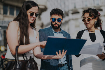 Canvas Print - Three young professionals wearing sunglasses review business documents outdoors on a sunny day. The group appears focused and engaged, highlighting teamwork, collaboration, and modern business attire.