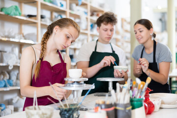 Wall Mural - Young woman teacher in apron teaches teenage boy and girl students to make ceramic cup in ceramic workshop