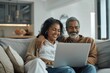 © zong - Middle aged couple smiling on sofa using laptop at home