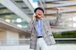 © Prostock-studio - Asian man wearing a stylish blazer and light pants expresses excitement while talking on the phone outdoors, celebrating a personal achievement in a modern urban environment.