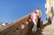 © Prostock-studio - A cheerful couple stands side by side on stone stairs, basking in the sun under a clear blue sky. They share smiles, dressed casually for a leisurely day outdoors in the city.