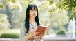 © Tati - Asian woman reading book outdoors in park, surrounded by greenery and sunlight