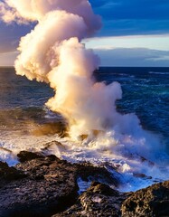 Wall Mural - Ocean waves crash against a rocky shore as a plume of steam rises from the water. Dramatic sunset colors light the scene.