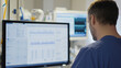 © pattozher - Intensive Care Monitoring: A focused male doctor meticulously reviews patient data on a large computer monitor in a modern hospital setting.  The image conveys a sense of urgency and precision.