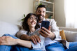 © Daniel - Happy caucasian couple relaxing at home using phone together looking social media.