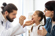 © sofiko14 - Caucasian doctor examines young girl's throat using wooden spatula. Scene takes place in a well-lit medical office, highlighting attentive interaction and professional healthcare.