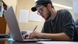 © Jiraphat - A focused young man works on a laptop, taking notes in a study environment, emphasizing productivity and learning.