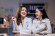 © Apichat - Two women are sitting at a desk, smiling and looking at each other