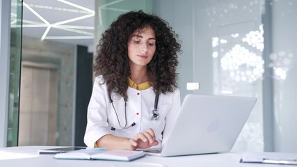 Focused young female doctor is using, typing on a laptop while sitting at a desk at a workplace in hospital clinic. A brunette woman medical worker physician works on a computer in the modern office