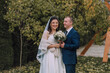 © Vasil - A bride and groom are posing for a picture in a park. The bride is wearing a white dress and a veil, while the groom is wearing a blue suit. They are both smiling and holding a bouquet and a vase