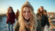 ©  Shomixer - A cheerful young woman, adorned in a knit hat and scarf, stands smiling in the foreground on a beach, celebrating friendship and happiness with her friends.