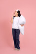 © PBXStudio - Full body shot of tired asian woman in white nightgown and eye mask hugging pillow and yawning covering mouth with hand, standing on pink background
