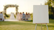 © Thamrong - A blank wedding welcome sign mockup on a contemporary metal easel, placed on a grassy field under soft natural lighting. The background features gently blurred wedding decor, including a floral arch