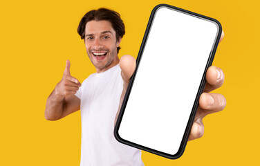 Poster - Cheerful young guy holding smartphone with white empty screen, pointing at blank display for mobile application advertisement or your text, gadget mockup template, isolated orange studio background