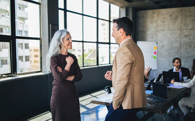 Fototapete - A man and a woman engage in a lively conversation in a bright, open office space. Their expressions suggest collaboration and creativity, with a team visible in the background.