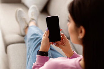 Poster - Over The Shoulder View Of Unrecognizable Young Woman Holding And Using Cell Phone, Sitting And Resting On The Couch In Living Room At Home. Selective Focus On Gadget, Template