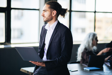 Aufkleber - A man in a suit stands with a laptop in hand, engaged in a meeting within a bright, contemporary office space. His colleagues are interacting in the background.
