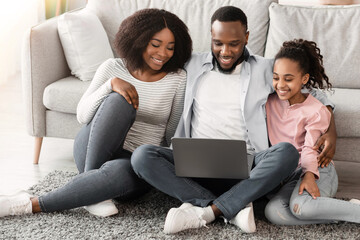 Wall Mural - Family Weekend. Cheerful African American parents and their little daughter using laptop at home together, watching movie or browsing internet, sitting on the floor rug carpet in living room, hugging