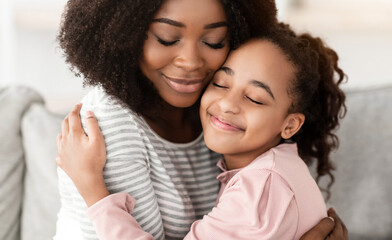 Wall Mural - Family Love And Tenderness. Closeup portrait of happy black woman with curly hair embracing cute little girl with closed eyes, spending free time together. Smiling mother and daughter hugging at home