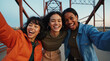 © Sachin - Three diverse young women friends laughing and taking selfie on bridge, multicultural Gen Z friendship, authentic joyful moment in casual winter outerwear