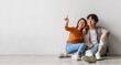 © Prostock-studio - Carefree millennial Asian man and woman sitting on floor near white studio wall and pointing at copy space, banner design. Young couple offering place for cool advertisement, panorama