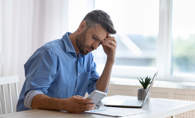 Poster - Worried male freelancer looking at smartphone screen while working at home office, received bad news, got scam message, thoughtful man touching forehead with concerned face expression, copy space