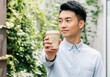 © Alexandra - Young asian man showing a coffee cup outdoors with a green background
