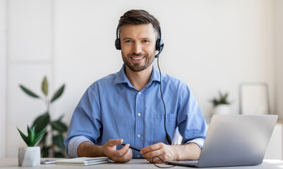 Poster - Portrait of smiling male customer service operator in headset posing at workplace, looking at camera, portrait of handsome hotline manager sitting at desk with laptop in white office, copy space