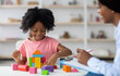 © Prostock-studio - Female child development specialist observing cheerful child preschooler, happy little african american girl sitting at table and making pyramids from colorful wood blocks. Child mental health concept