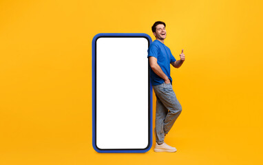 Poster - Cool Mobile Offer. Excited casual guy leaning on big smartphone with blank empty white display, showing thumb up gesture, recommending great new app or website, mock up, full body length, yellow wall