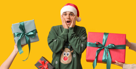 Sticker - Surprised young man in Santa hat receiving Christmas gifts on yellow background