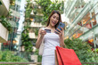 © StockPhotoRepublic - Asian Woman In Casual Attire Carrying Shopping Bag Using Phone s and Credit Card Happily Amid A Modern Shopping Mall Building with Greenery Outdoors