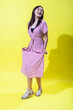 © Daniel - Asian woman in a pink floral dress holding its hem playfully, smiling warmly and posing confidently against a vibrant yellow background, showcasing a cheerful and stylish summer look.