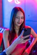 © StockPhotoRepublic - Happy Young Asian Woman Using Phone in a Neon-Lit Lounge with Shopping Bags