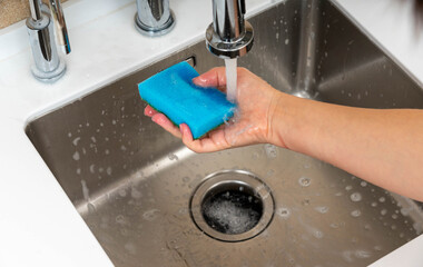 A close-up of a blue sponge being washed under running water in a stainless steel kitchen sink, ensuring cleanliness and hygiene.