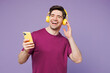 © ViDi Studio - Young man he wear violet t-shirt casual clothes listen to music in headphones hold use mobile cell phone record voice sing isolated on plain pastel light purple background studio. Lifestyle concept.