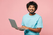© ViDi Studio - Young smiling happy IT Indian man he wear blue t-shirt casual clothes hold use work on laptop pc computer look camera isolated on plain pastel light pink background studio portrait. Lifestyle concept.