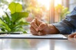 © Vilaysack - Hand holding a pen, signing a document on a desk in a well-lit office with green plants and natural sunlight.