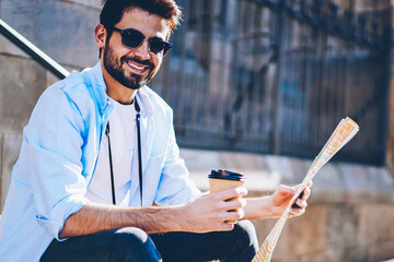 Wall Mural - Cheerfu bearded traveler happy with trip in old town on weekends holding coffee to go in hand while smiling at camera.Positive stylish tourist in sunglasses with travel map sitting on street stairs