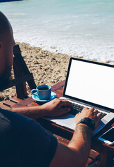 Fototapete - Back view of skilled male freelancer doing distance job on ocean shore browsing information on laptop computer,man travel blogger typing post for update content on website recreating on sea beach