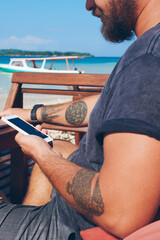 Fototapete - Cropped image of man holding mobile phone checking banking balance and making transactions sitting on ocean beach, male freelancer using technology during remote job synchronize cellphone and netbook