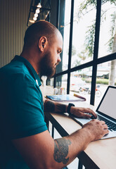 Fototapete - Young bearded guy sitting in cafeteria with laptop computer typing text for publication in blog,skilled male freelancer doing distance job via netbook with mock up screen and wireless internet