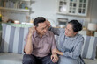 © ultramansk - An elderly couple sits on a couch in a cozy living room. The man holds his head in discomfort, while his partner offers support. The image highlights care, concern, and the bond between the couple.