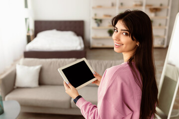 Poster - Portrait of smiling young woman holding digital tablet with black empty screen for mockup template, looking back at camera, standing in modern living room. Technology, Automation, Smart Home Concept