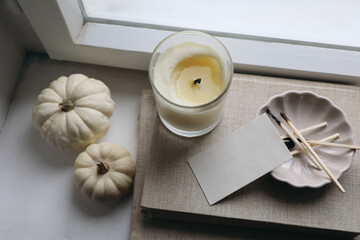 Wall Mural - Moody autumn, winter still life. Candle in glass, old books. Matches, ceramic plate on windowsill. Blurred white pumpkins. Blank business card, tag mockup. Fall Thanksgiving. Halloween. Top view.