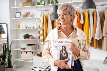 Wall Mural - Happy mature female stylist with magazines against stylish clothes in studio